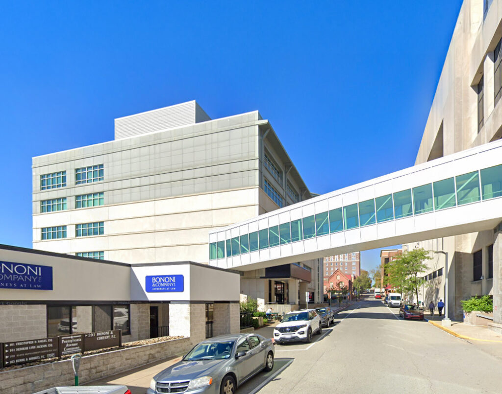 An elevated glass pedestrian bridge connecting two buildings over a street with parked cars and a law firm sign in the foreground.