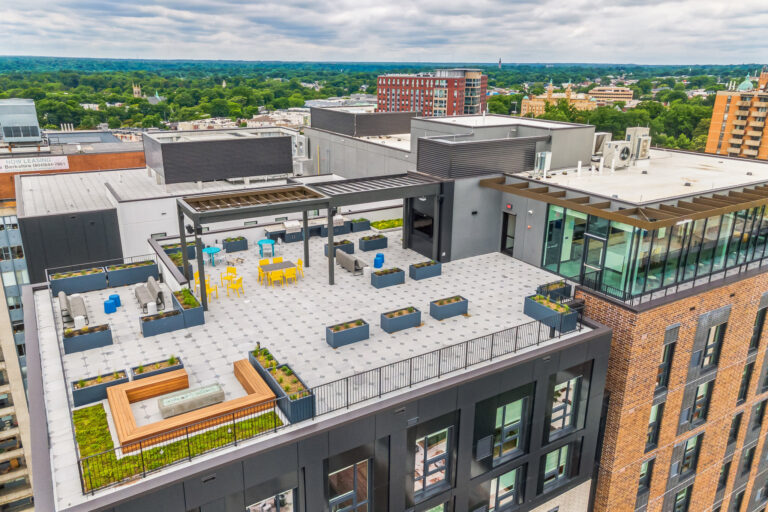 Aerial view of a modern apartment building rooftop terrace featuring lounge seating, dining tables, and potted planters.