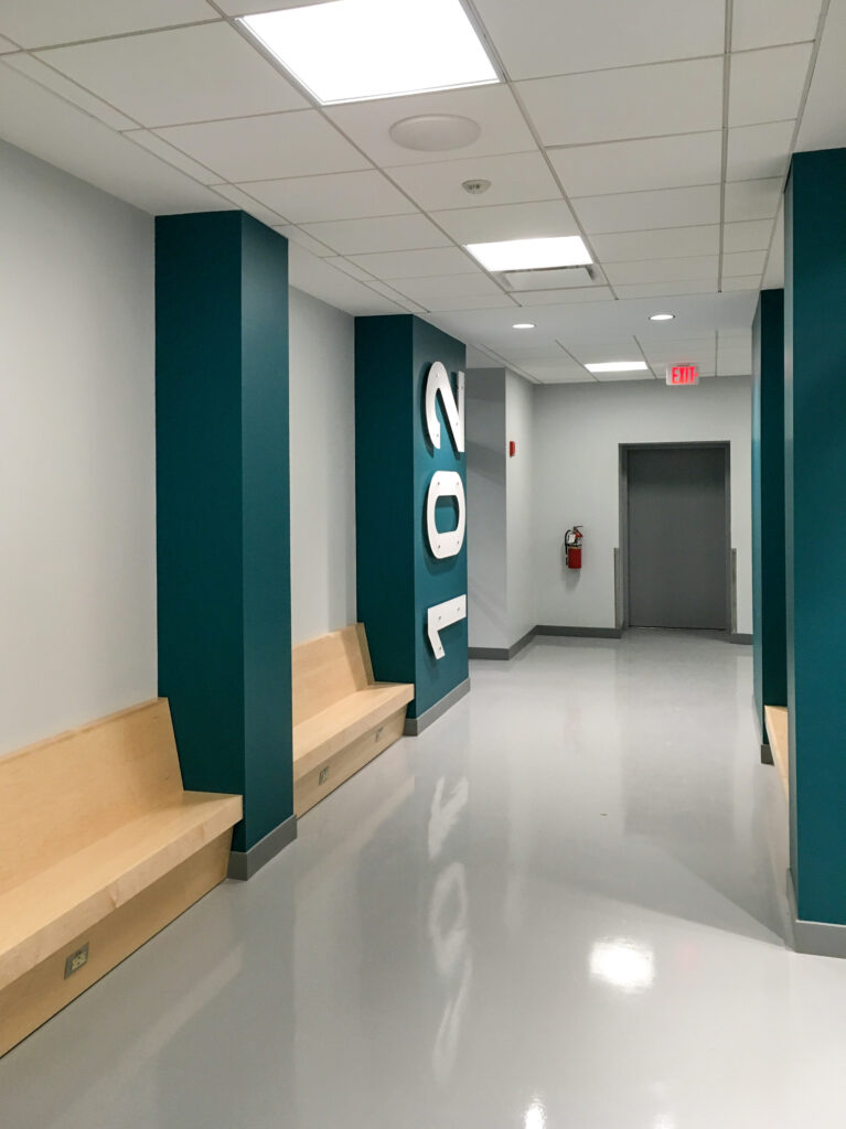 A hallway with light wood benches along teal support columns and a large number 102 mounted on the wall.