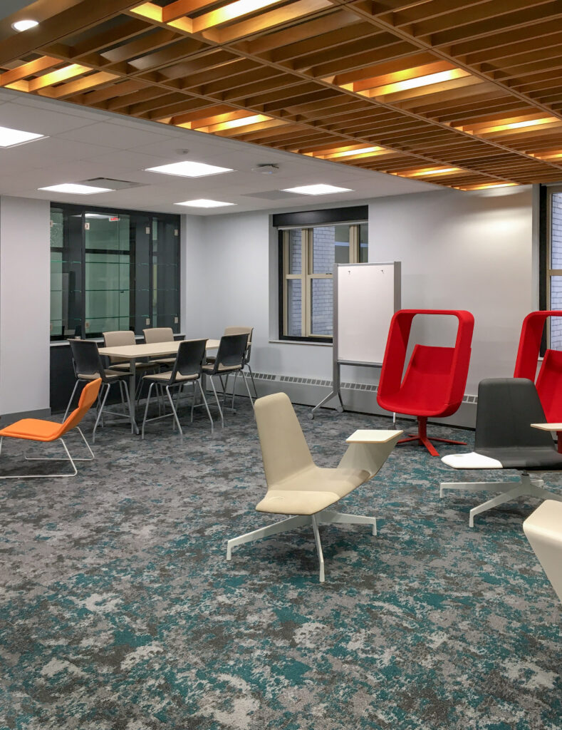 A modern office lounge with a wooden slat ceiling, a conference table with chairs, and various standalone chairs including red enclosed seating.