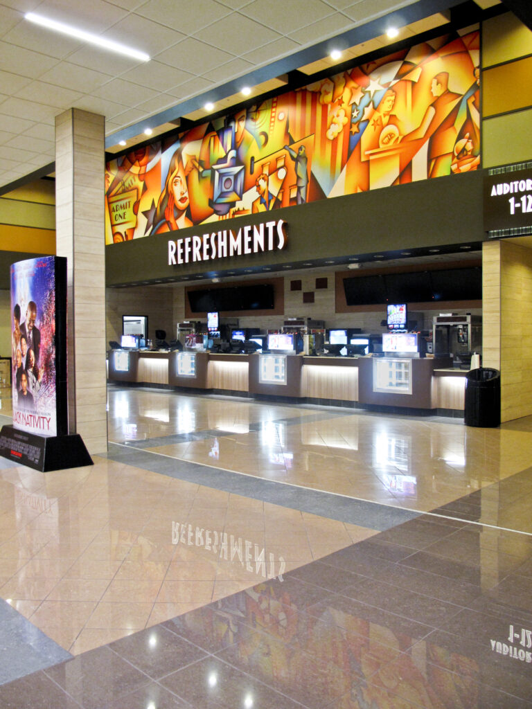 Movie theater concession stand with a “REFRESHMENTS” sign and a colorful mural above, and a movie poster for “Black Nativity” on a stand in the foreground.