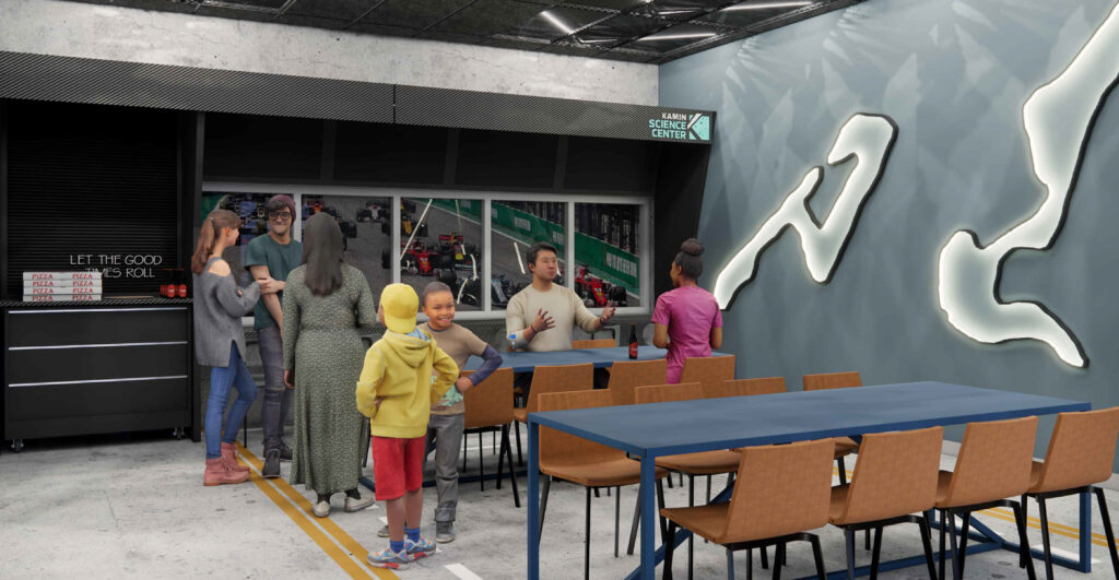 A group of people standing and sitting in a modern room with a Kamin Science Center sign, a race car mural, and a glowing wall graphic.