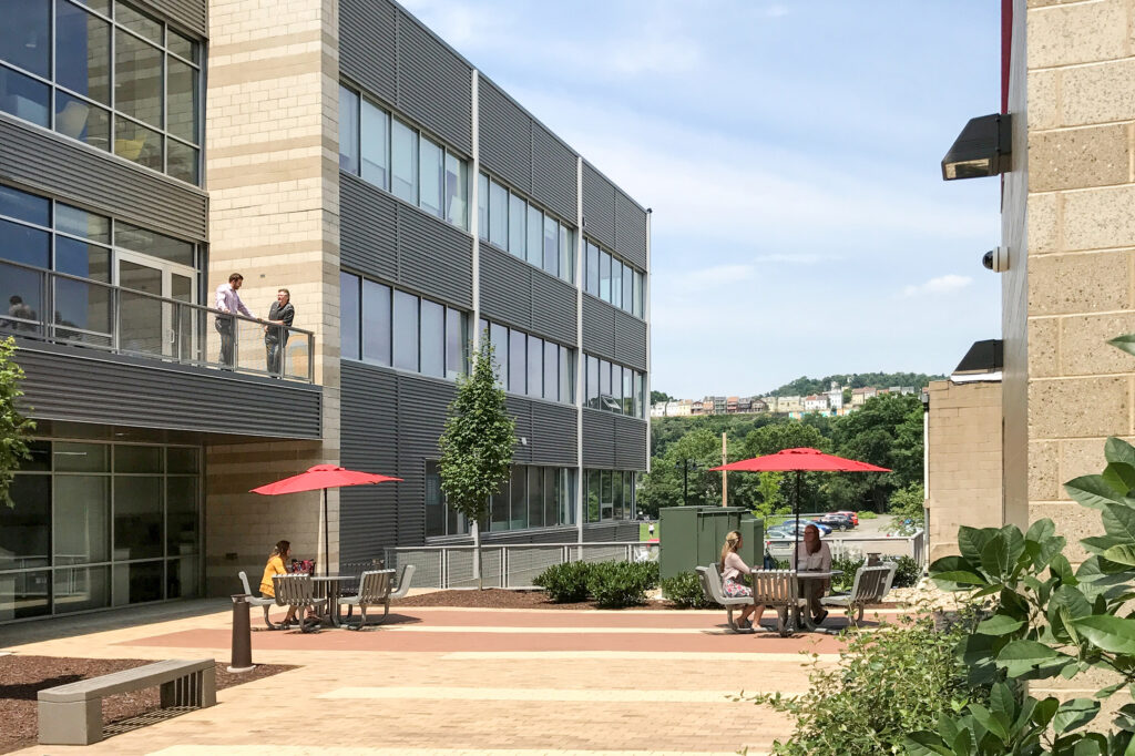 People seated at outdoor tables under red umbrellas by a modern office building, with two people standing on a second-floor balcony above.
