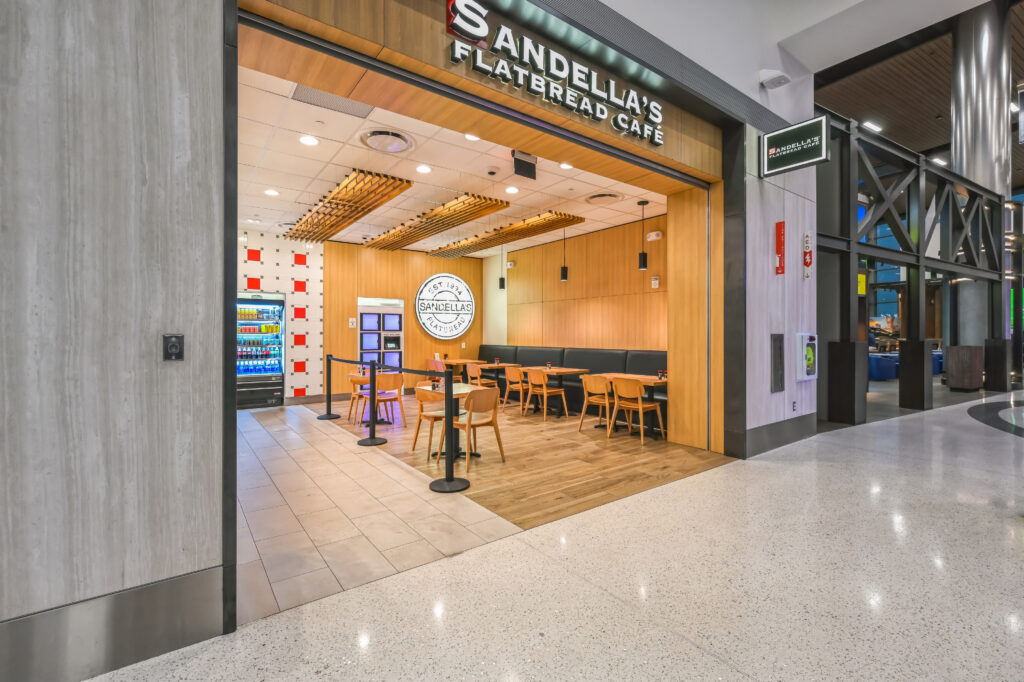 Sandella’s Flatbread Cafe storefront in a shopping center, featuring an open dining area with tables, chairs, and a refrigerated display case.