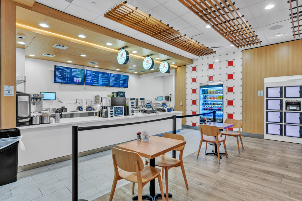 Interior of a cafe with a service counter, digital menu boards, and wooden tables and chairs.