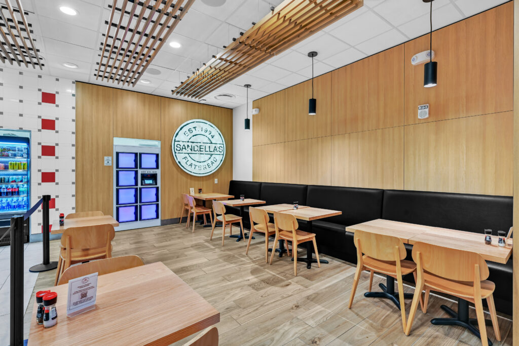 Interior of a Sandella’s Flatbread restaurant with wooden tables and benches, and a circular wall sign featuring the brand logo.