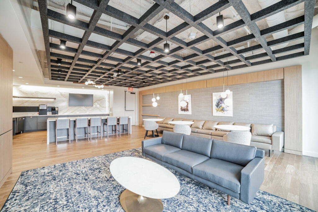 Modern office lounge area with a dark grey sofa, a white oval coffee table, a long bench seat with small tables, and a kitchenette with bar seating under a dark grid ceiling.