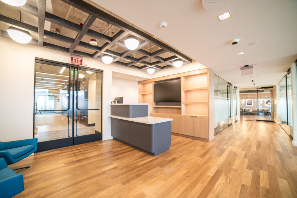 Modern office reception area with a gray front desk, wooden shelving, glass-paned doors, and a grid-patterned ceiling.