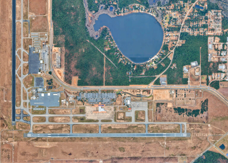 Aerial view of an airport terminal and runway situated next to a large lake and wooded residential area.