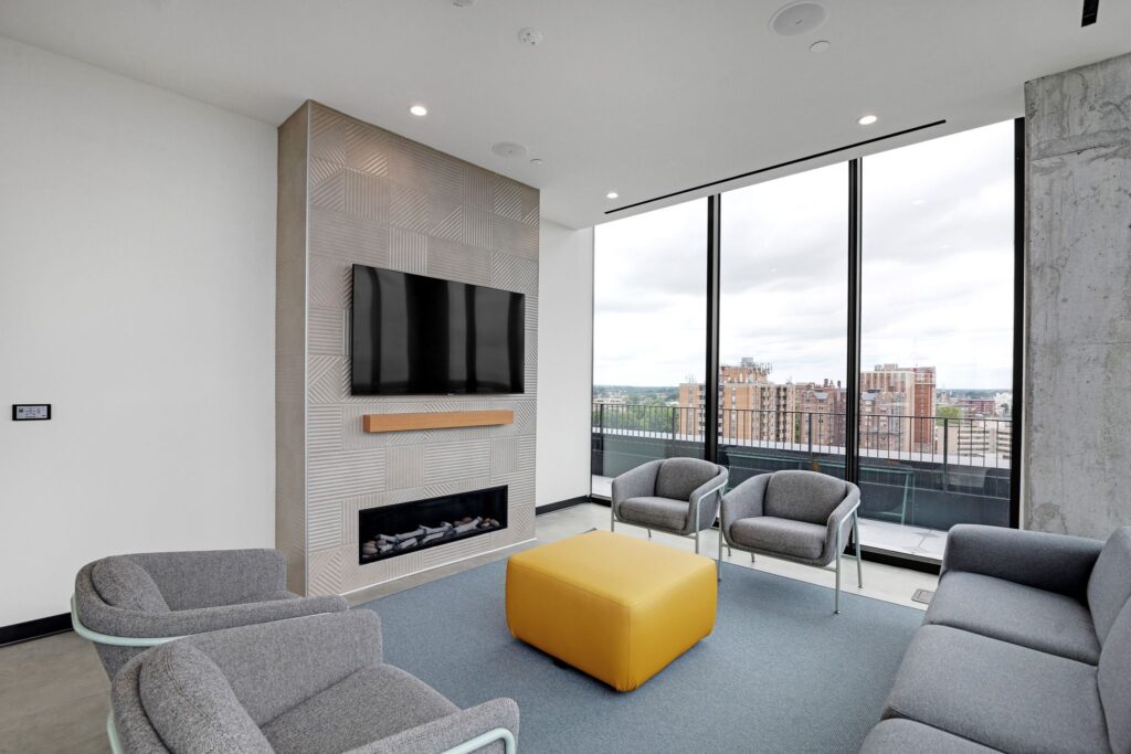 A modern lounge area with gray armchairs, a yellow ottoman, and a wall-mounted television above a linear fireplace, featuring large windows overlooking a city skyline.