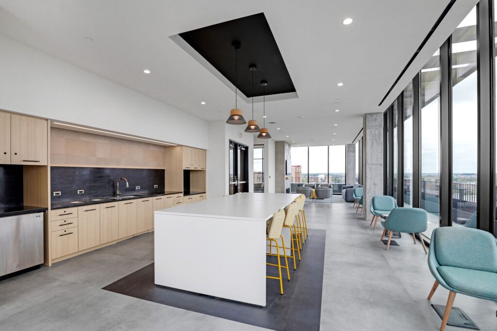Modern kitchen and lounge area featuring a large central island with bar stools, light wood cabinetry, and floor-to-ceiling windows overlooking a city skyline.