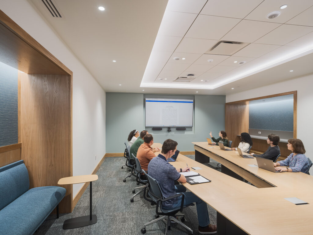 A group of people sit around a large conference table in a meeting room, facing a screen on the wall.