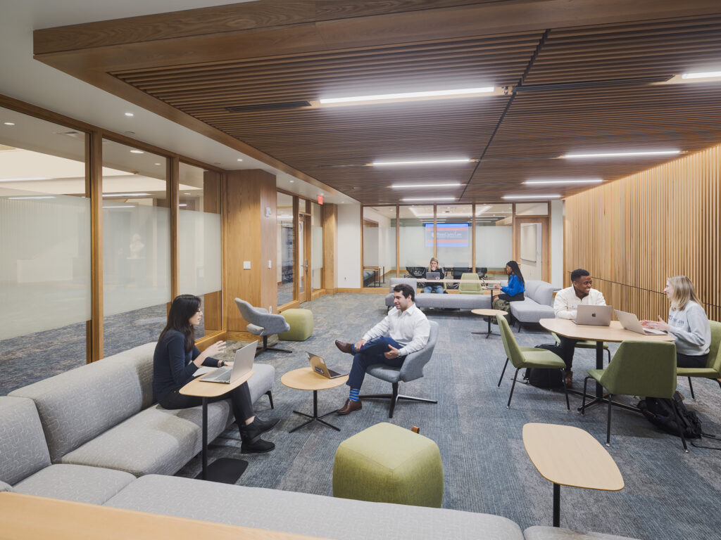 People working on laptops in a modern, open-plan office lounge with wooden slat ceilings and glass-walled conference rooms.