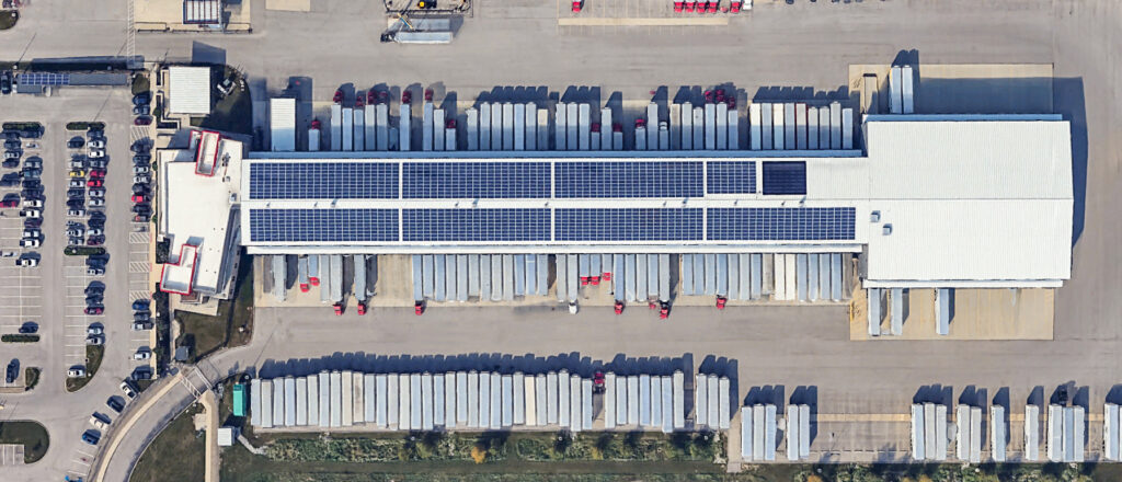 Aerial view of a large logistics warehouse with solar panels on the roof and rows of parked semi-trailers in the surrounding shipping yard.