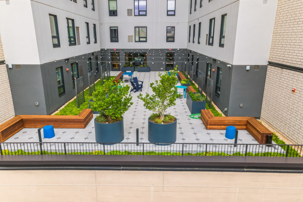 An aerial view of an apartment complex courtyard featuring two large potted trees, wood bench seating, and chairs arranged on a patterned patio.