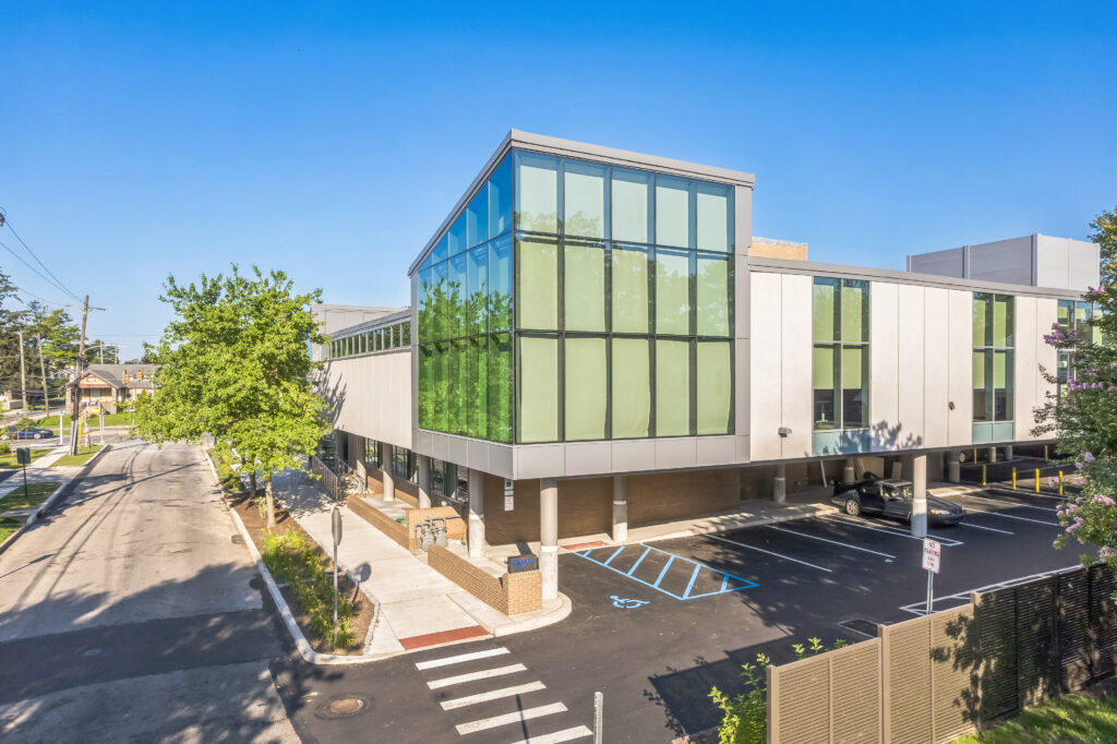 Modern two-story office building with a glass corner facade, parking lot, and pedestrian crosswalk.