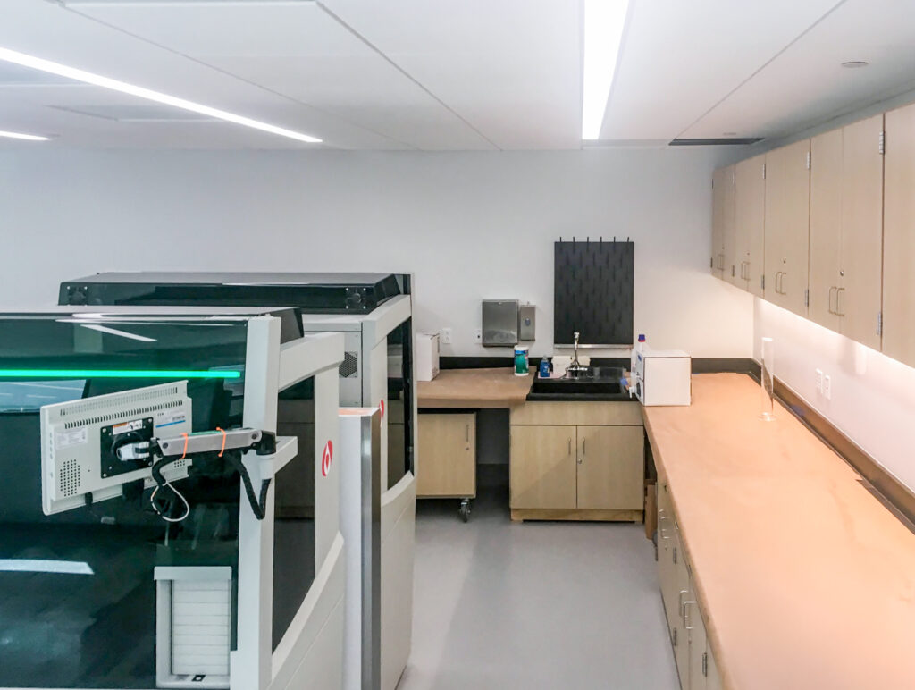 A laboratory interior with large analytical equipment in the foreground and wooden cabinetry and a sink along the back wall.