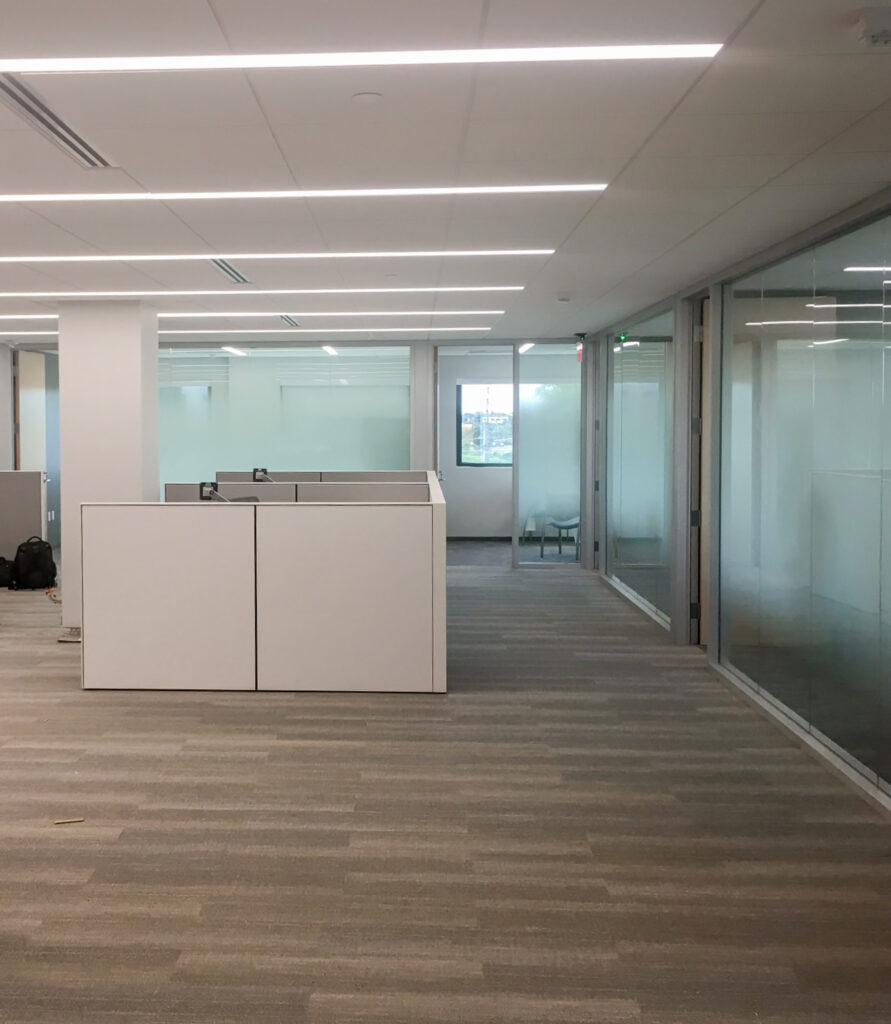 An empty modern office featuring open-plan cubicles, glass-walled conference rooms, and gray carpeted flooring.
