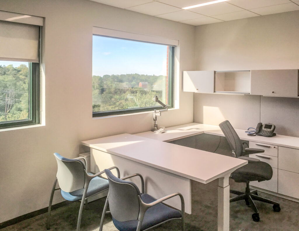 An office with a white L-shaped desk, a rolling office chair, two guest chairs, and a telephone, facing a large window with a view of trees.