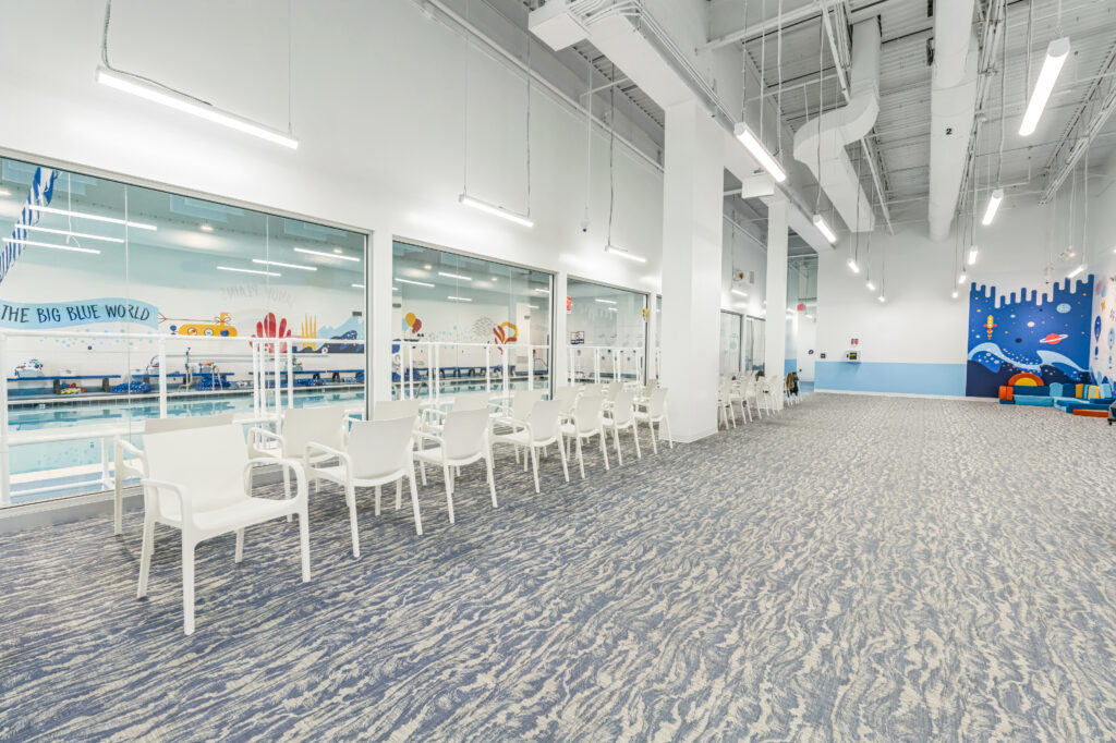 A row of white plastic chairs facing an indoor swimming pool, seen through a large glass wall in a room with blue patterned carpeting.
