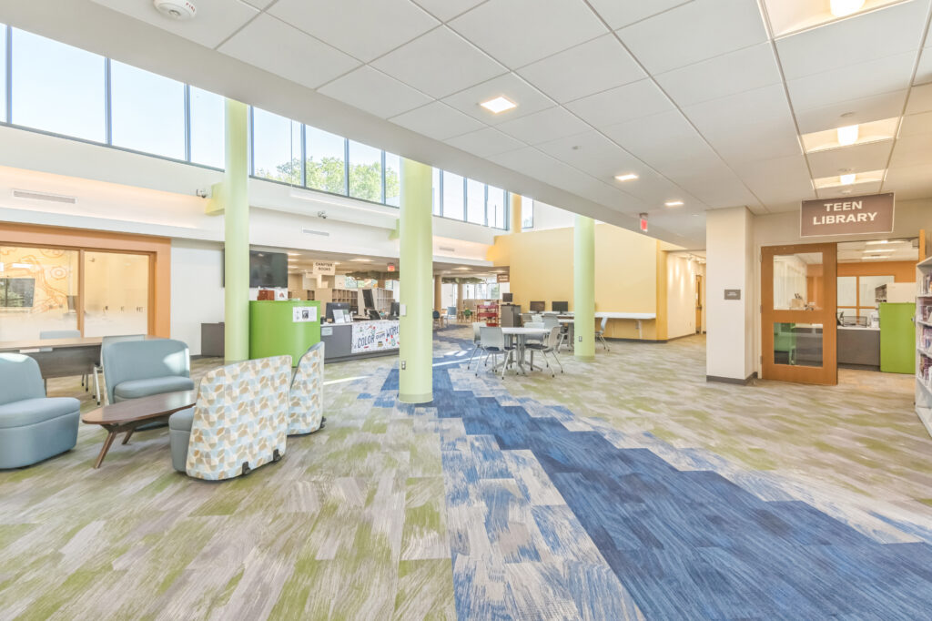 The interior of a library featuring a lounge area with armchairs, a service desk, computer workstations, and a doorway marked “Teen Library.”