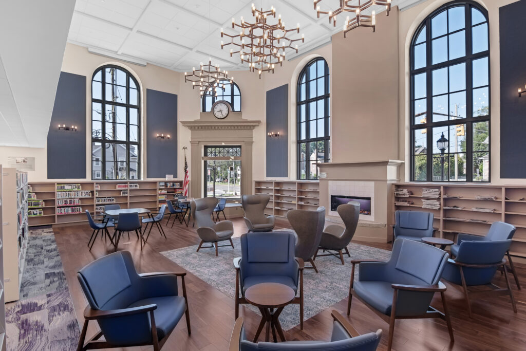 Library lounge area with blue armchairs, a central fireplace, arched windows, and wall-to-wall bookshelves.