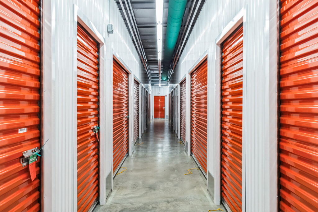 A long hallway in a storage facility lined with closed orange roll-up doors on both sides.