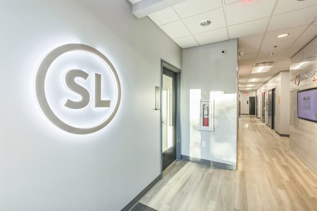 A hallway in a Salon Lofts location, featuring a backlit “SL” circular logo on the wall and several studio doors.