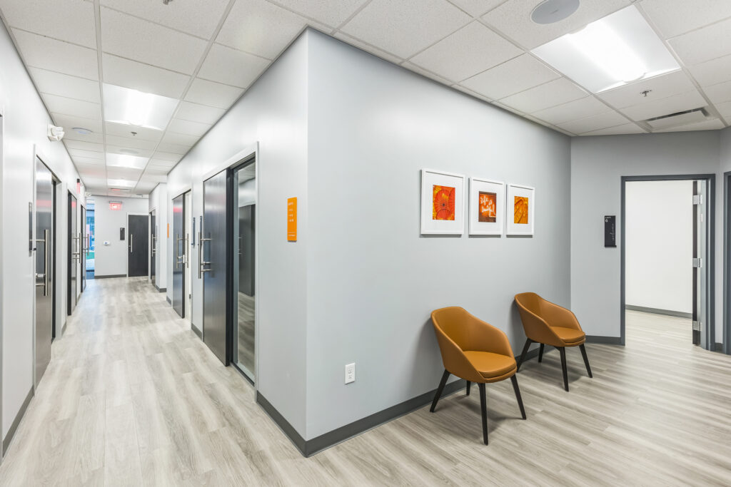 Office hallway with light wood flooring, several doors, and two tan chairs beneath framed art.