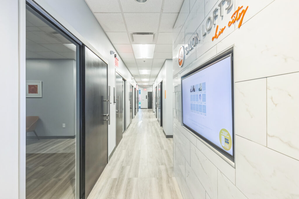 A brightly lit hallway in a Salon Lofts location, featuring dark doors on one side and a digital directory mounted on a marble-tiled wall.