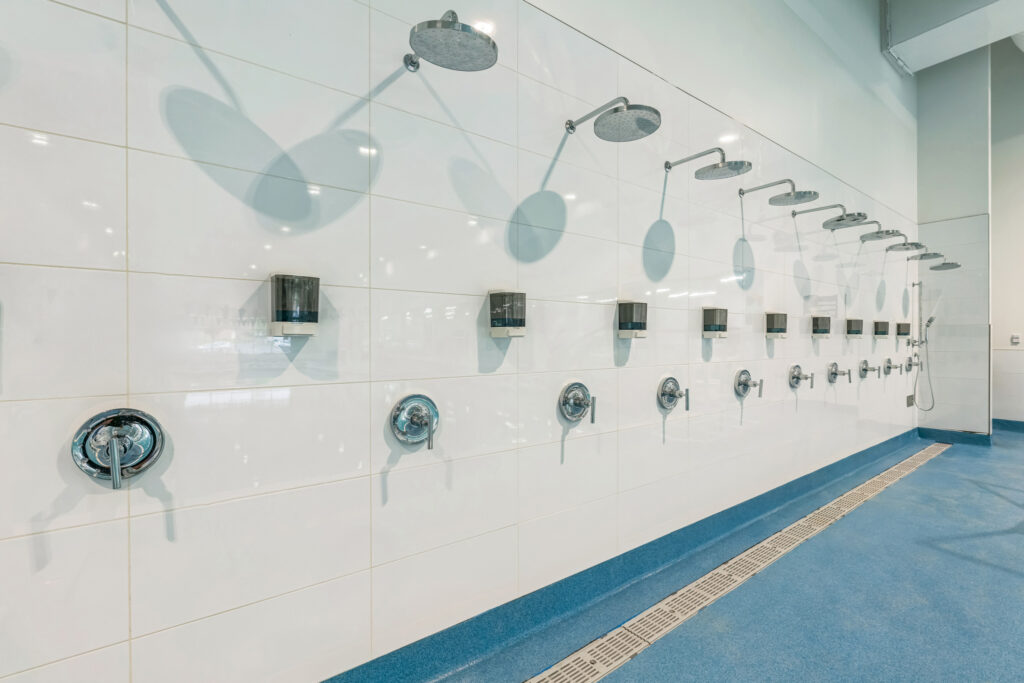 A row of shower heads and soap dispensers mounted on a white tiled wall above a blue floor with a drainage trench.