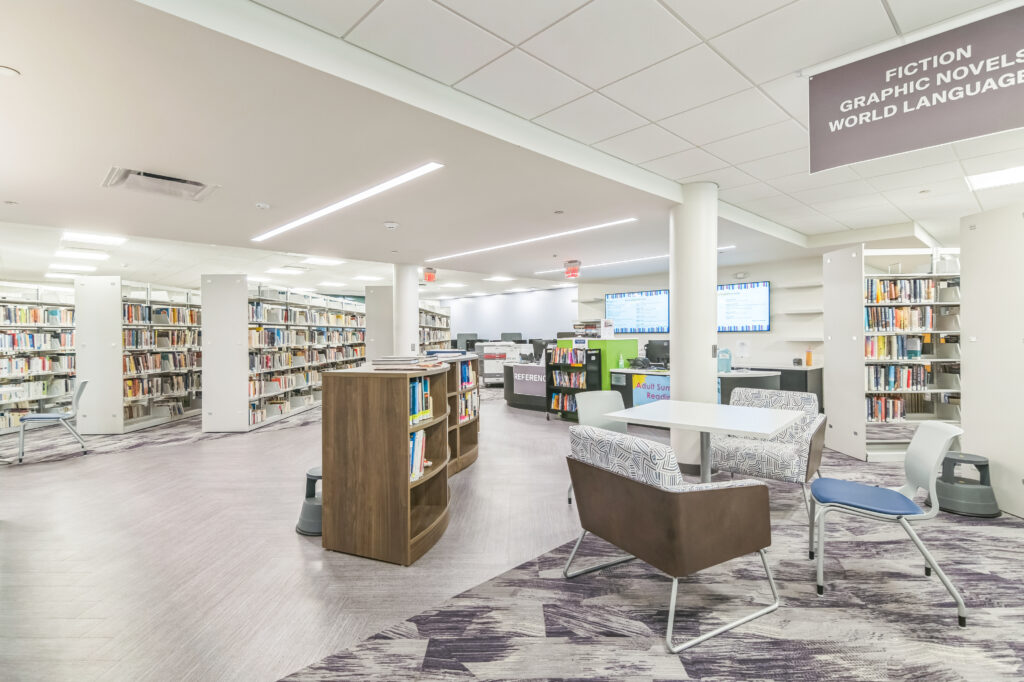 A modern library interior featuring rows of bookshelves, a central reference desk, and a seating area with chairs and a table.