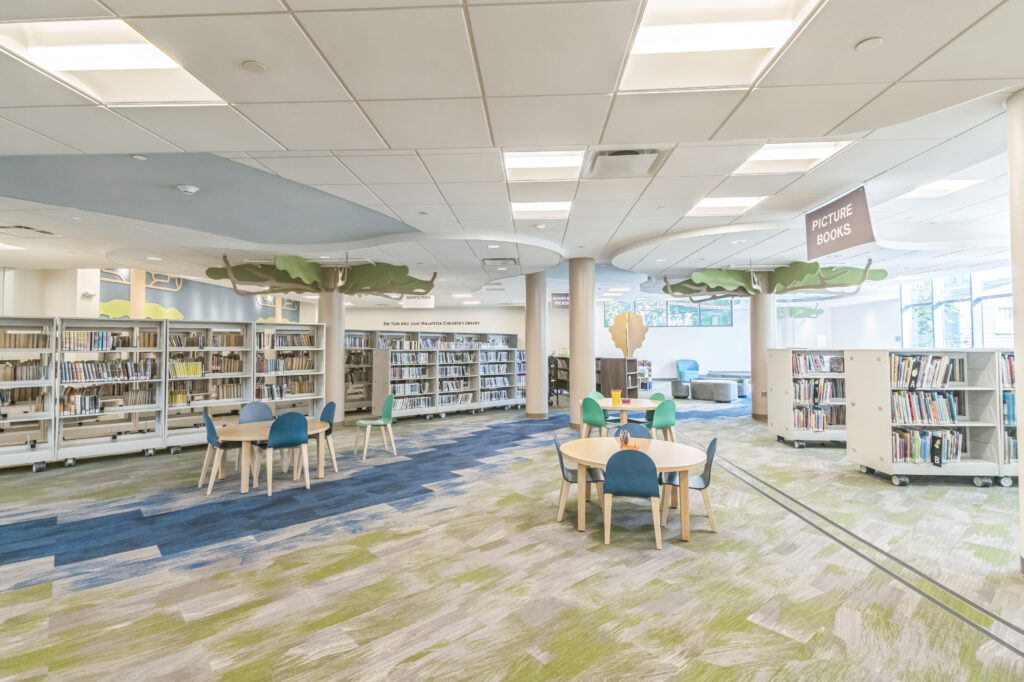 Children’s library section with rows of bookshelves, circular tables with blue chairs, and decorative tree cutouts on the ceiling.