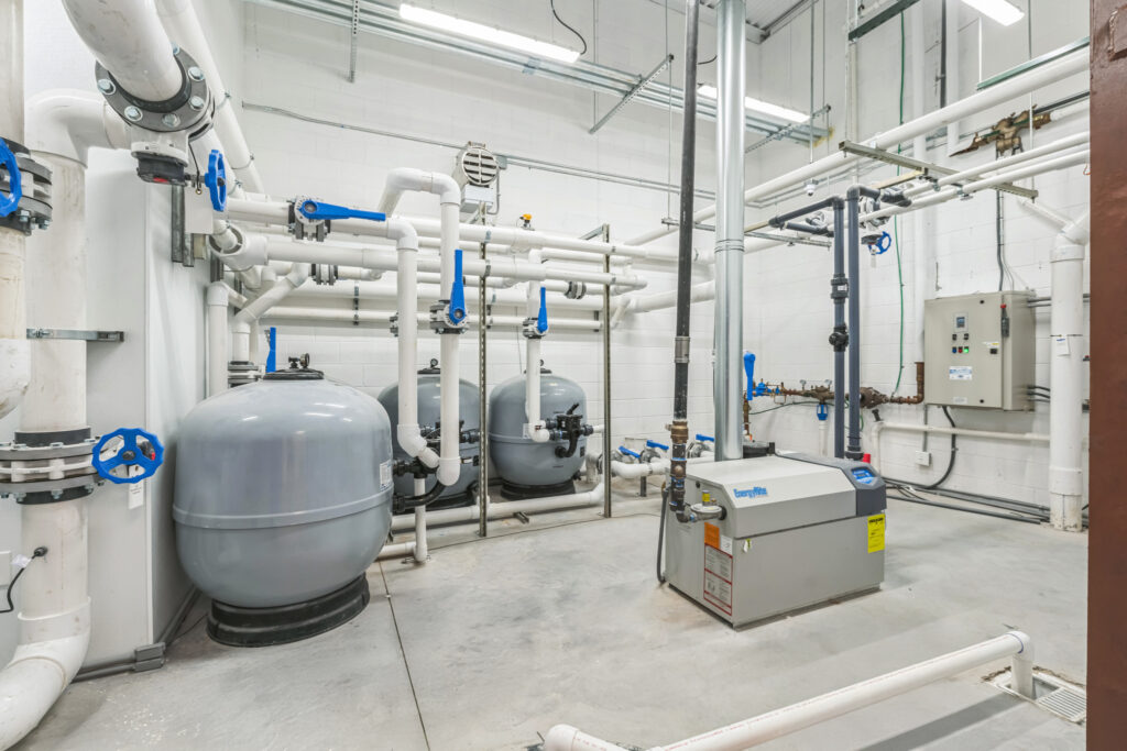 Mechanical room featuring two large grey pressurized tanks, white PVC piping with blue valves, and an EnergyRite pool heater.