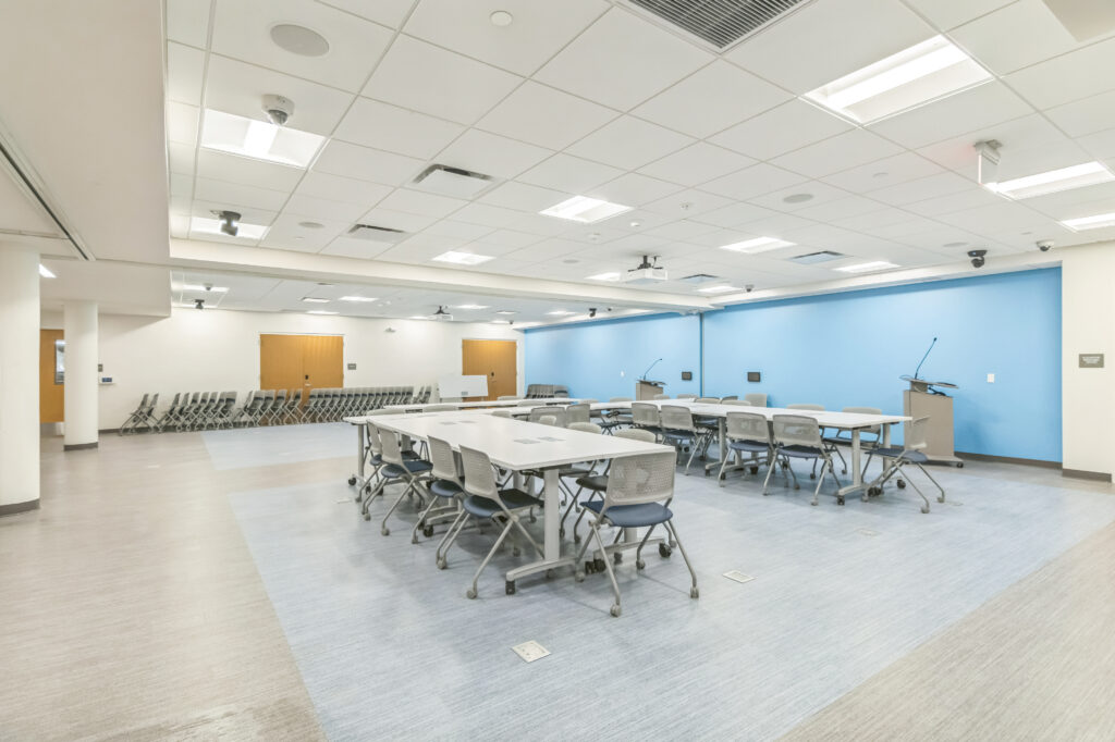 A large multipurpose room with rows of modular tables, rolling chairs, and two podiums against a blue accent wall.