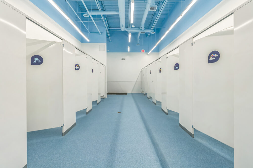 A hallway of white bathroom stalls with blue flooring and a blue exposed ceiling.