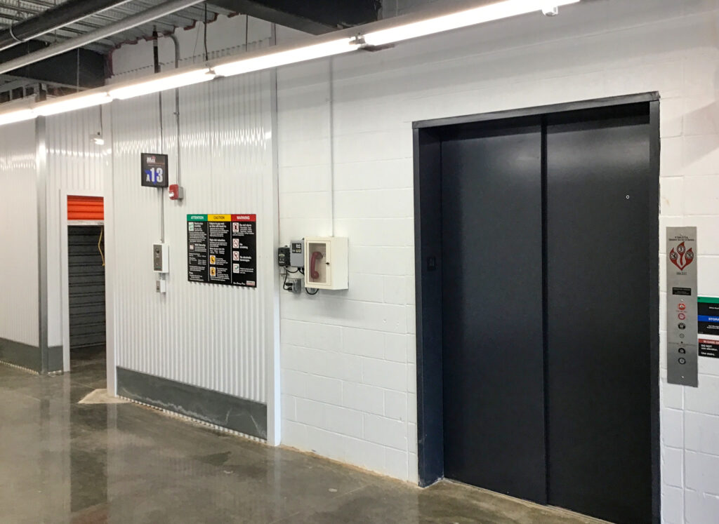 An elevator and adjacent storage unit door in a warehouse hallway with wall-mounted safety signs and an emergency telephone box.