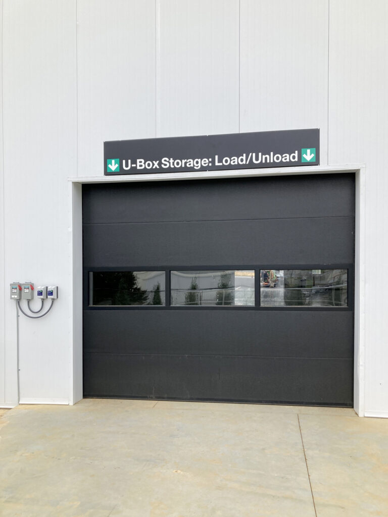 A black garage door with a row of windows and a sign above that reads “U-Box Storage: Load/Unload”.