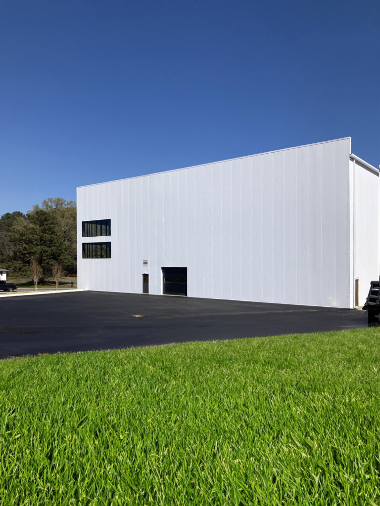 A large white industrial building with a flat, paved parking lot and a green lawn in the foreground.