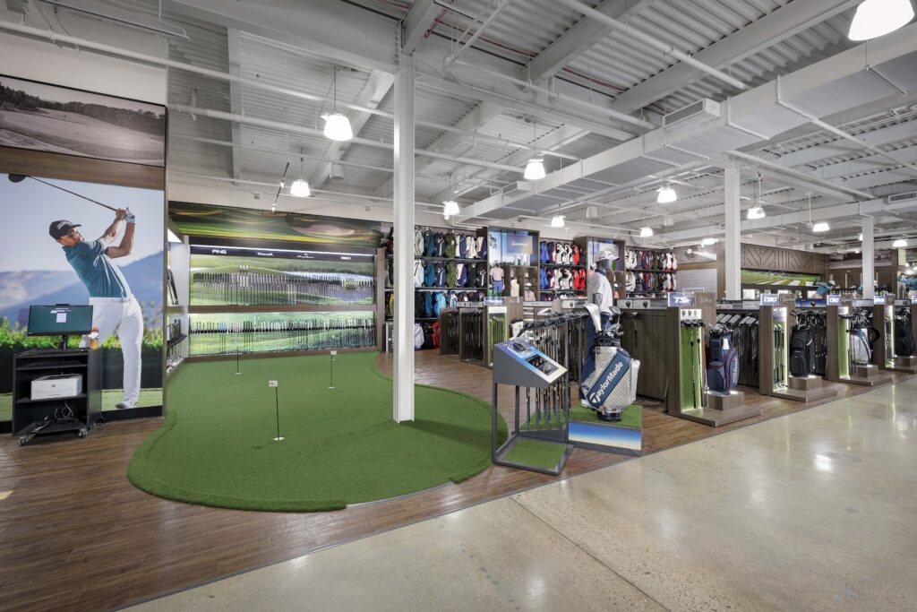 A golf retail store featuring an indoor putting green, golf club displays, and racks of golf bags.