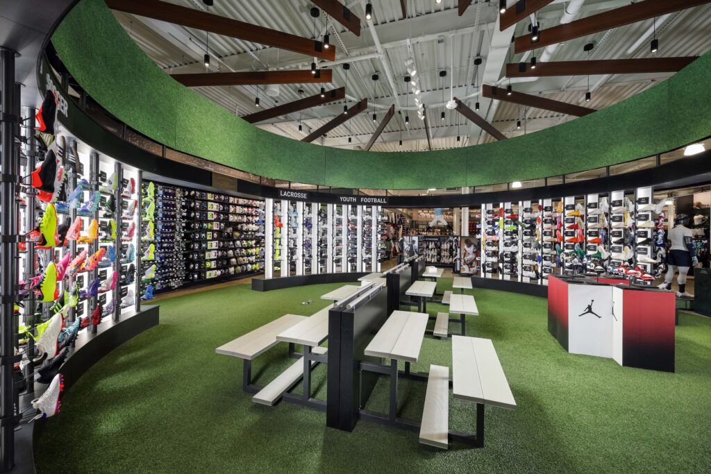 Interior of a sports store featuring rows of athletic footwear on display, artificial turf flooring, and central bench seating.