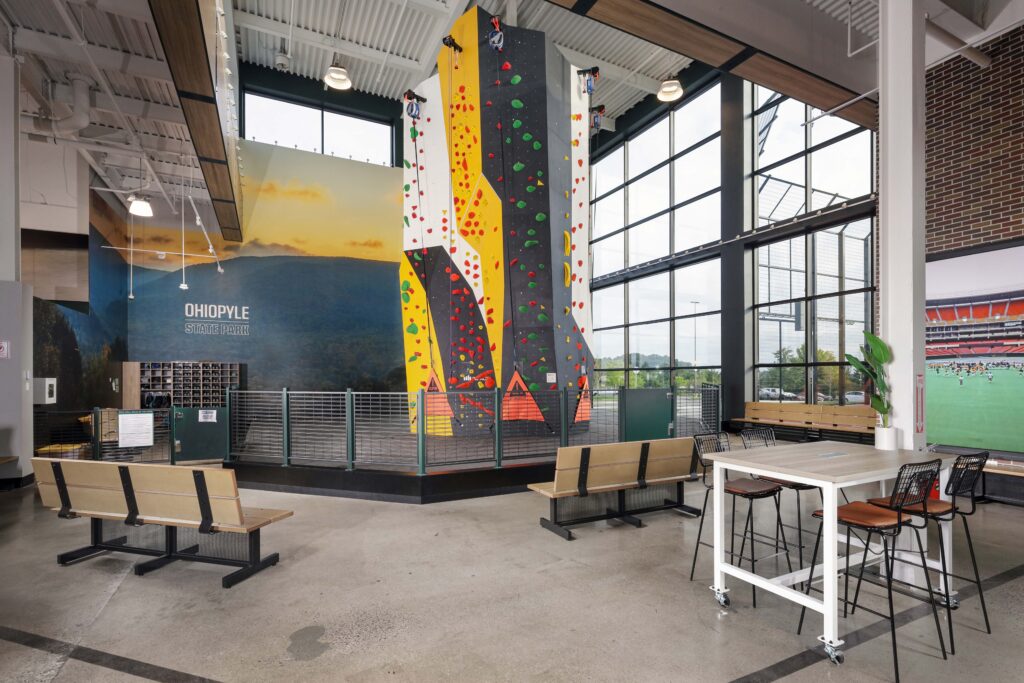 Indoor rock climbing wall with colorful holds situated in a facility featuring benches, a table, and a mural of Ohiopyle State Park.