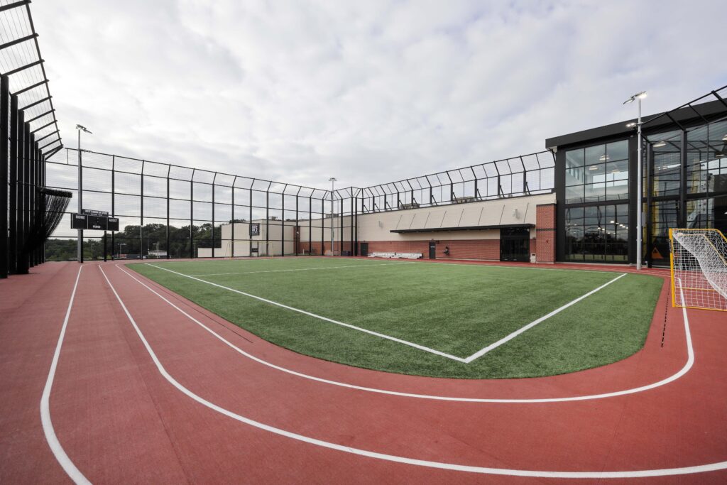 An outdoor facility featuring a red running track surrounding a rectangular artificial turf field, enclosed by a tall wire fence.