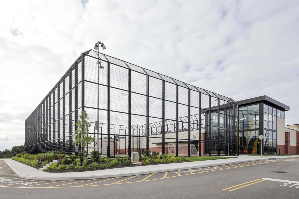 A modern sports complex featuring an outdoor court enclosed by tall black metal fencing, adjacent to a multi-story building with glass walls.