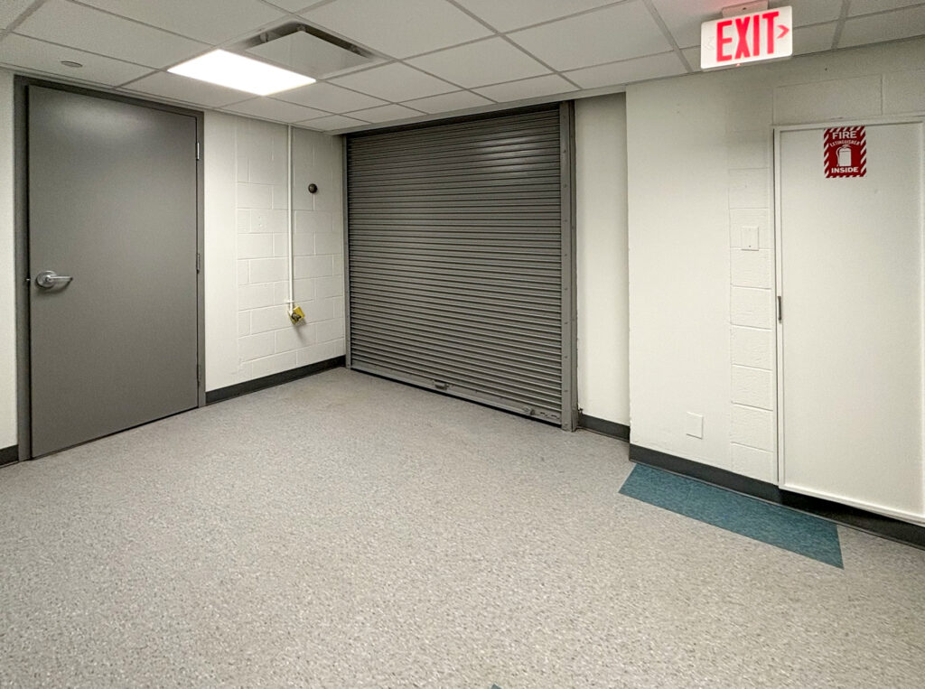 A room with a grey door, a closed metal rolling security gate, and a utility cabinet labeled “Fire Extinguisher Inside” under an illuminated exit sign.