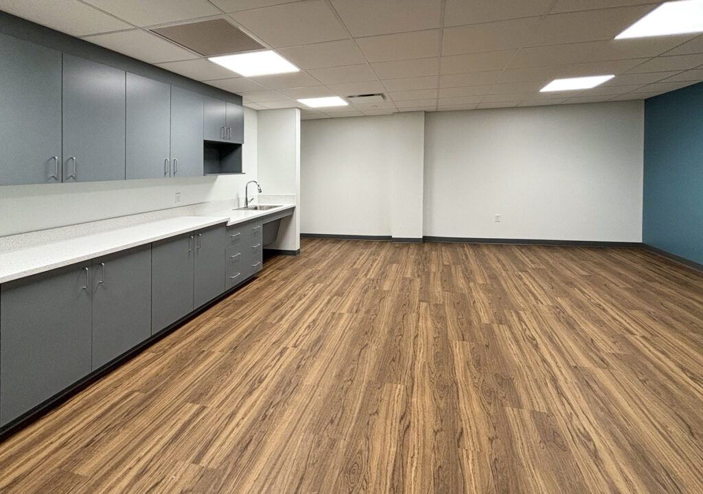 An empty office breakroom with grey cabinets, white countertops, a sink, and wood-patterned flooring.