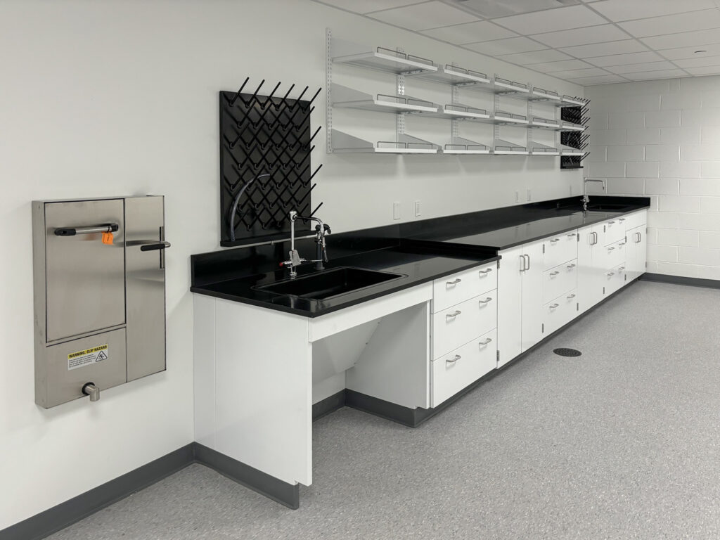 Laboratory workstation with white cabinets, a black countertop, a sink, wall-mounted drying racks, and a stainless steel wall-mounted cabinet with a warning label.