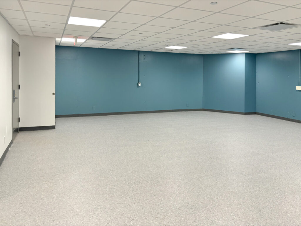 An empty room with teal walls, a light gray floor, and a drop ceiling with recessed lighting.