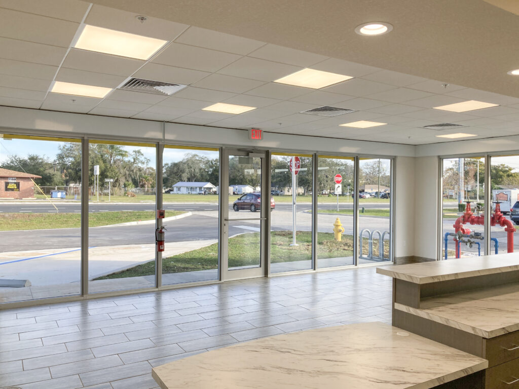 Interior view of an empty commercial space looking out through floor-to-ceiling windows toward a parking lot and street.