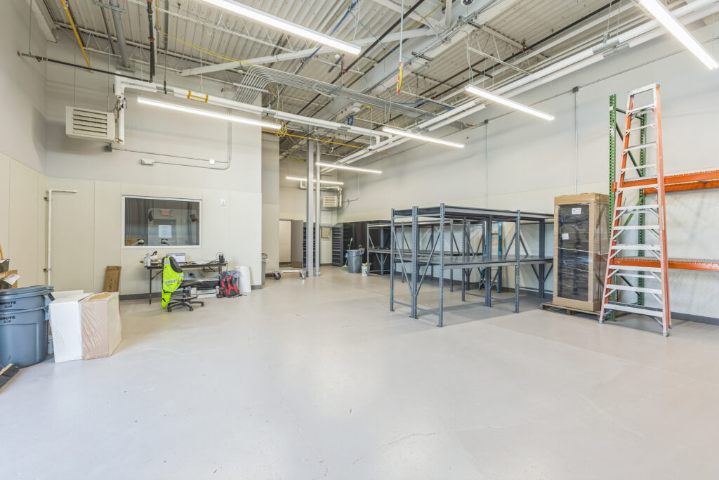 Interior of a warehouse space with empty metal shelving units, a desk with office equipment, and an orange step ladder leaning against a rack.